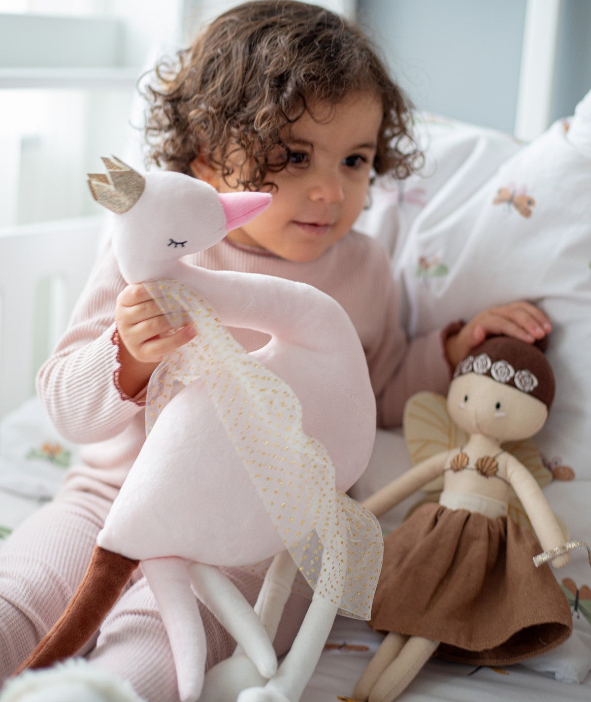 Criança com cabelo encaracolado a segurar o peluche de cisne Ivy ao lado de uma boneca de pano, em ambiente de brincadeira.