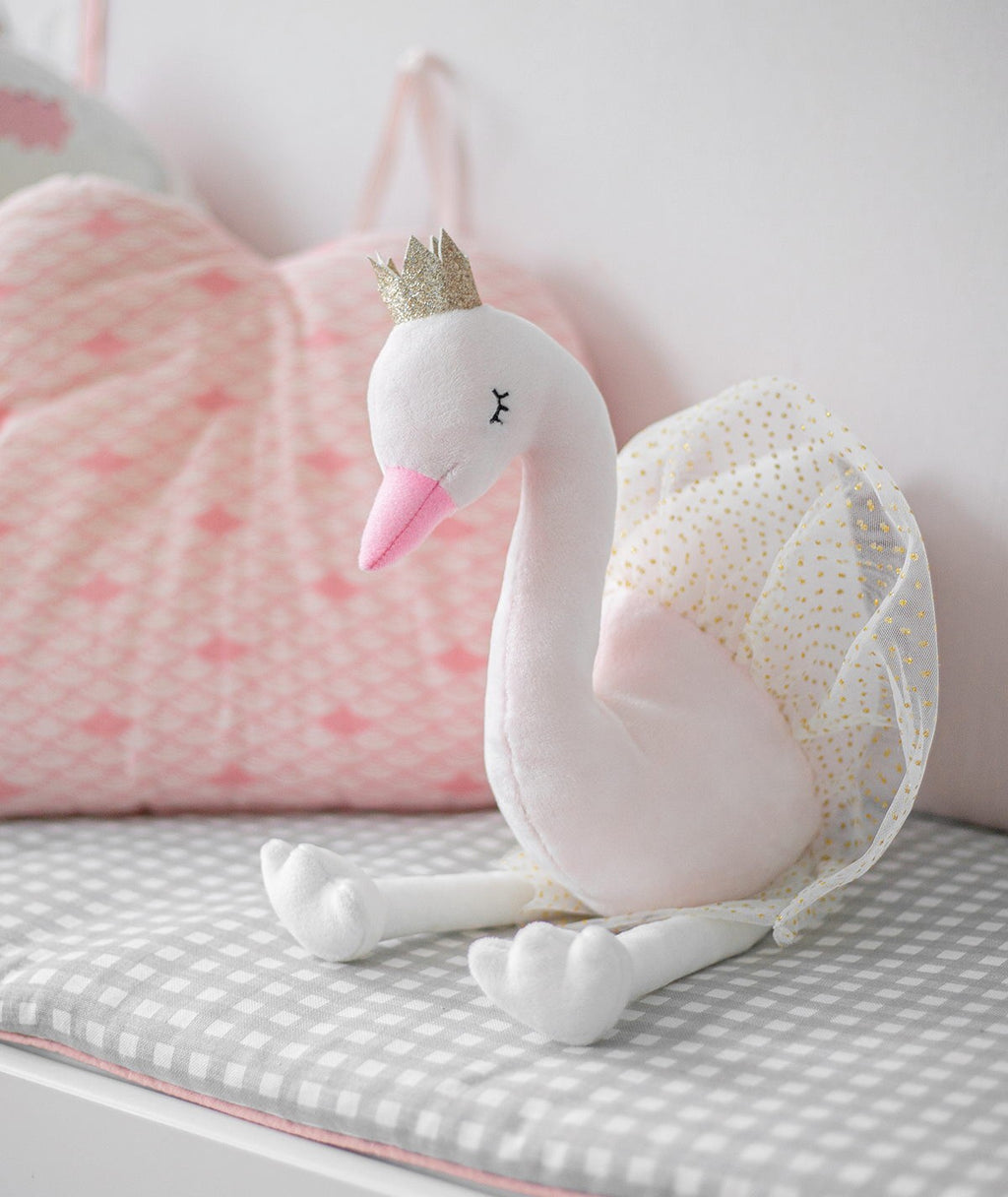 Peluche de cisne Ivy rosa sentado numa superfície xadrez cinzenta, com almofadas cor de rosa ao fundo, mostrando o seu tamanho e detalhes.