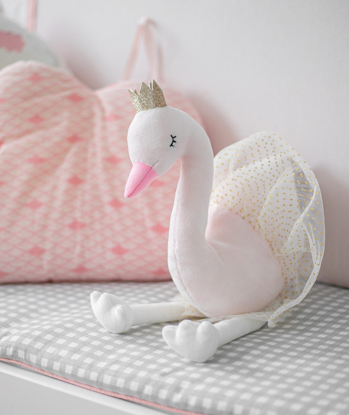 Peluche de cisne Ivy rosa sentado numa superfície xadrez cinzenta, com almofadas cor de rosa ao fundo, mostrando o seu tamanho e detalhes.
