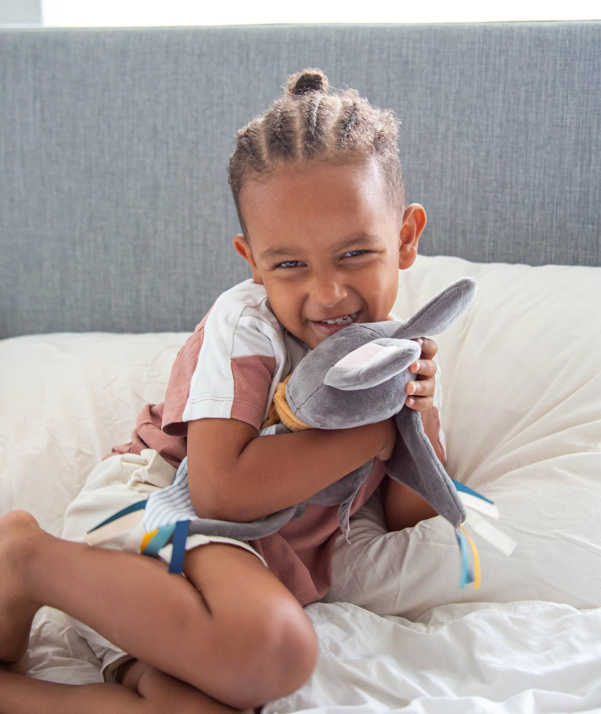 Criança sorridente abraça o peluche de morcego Bruce cinzento na cama, mostrando o uso do brinquedo em contexto de brincadeira.