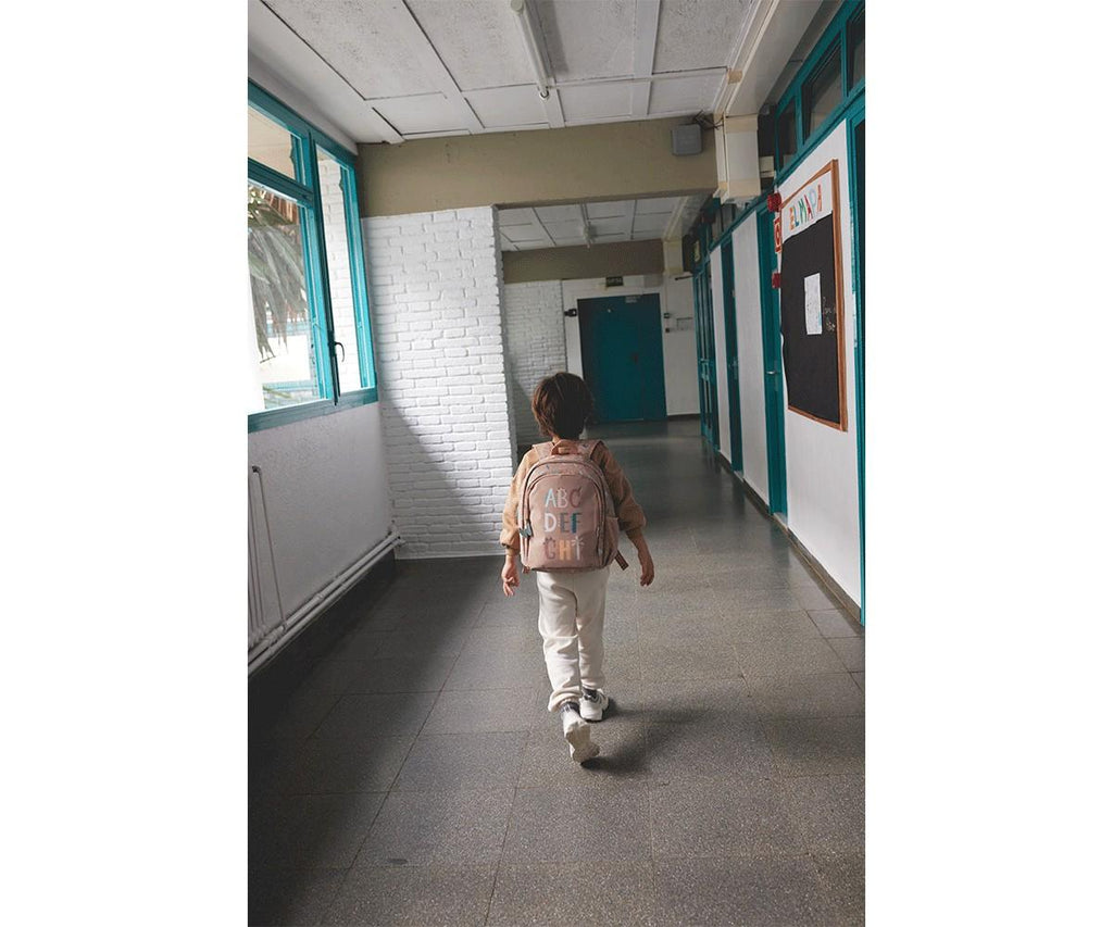 Criança de costas com a mochila Tutete Funny Letters bege a caminhar num corredor de escola.