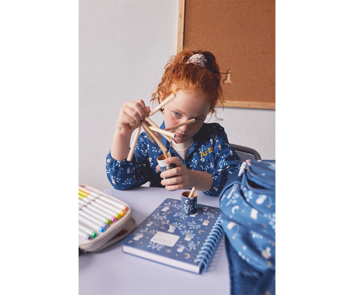 Criança de cabelo ruivo a usar o tubo de lápis Tutete para pegar lápis de cor de madeira, num ambiente de estudo com caderno e estojo.