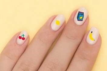 Detalhe de quatro unhas com autocolantes OMY 'Yummy' com desenhos de cerejas, um ponto amarelo, um pacote de batatas fritas e uma banana.