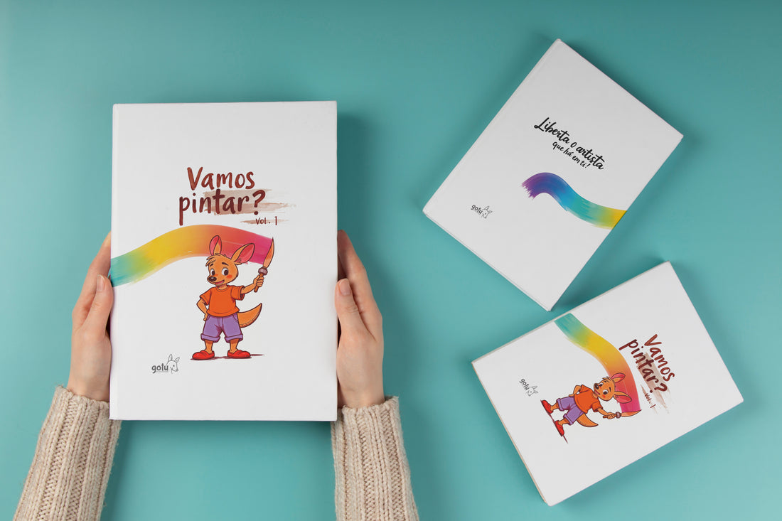 Mãos segurando o livro "Vamos Pintar? Vol. 1" com capa colorida e ilustração de um canguru pintando. Dois outros livros da mesma coleção aparecem ao lado.