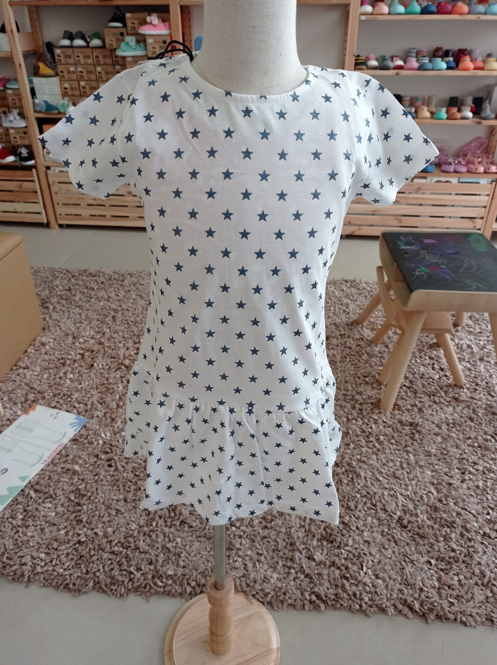 Vestido infantil Hublot branco com estrelas azuis, manga curta, em manequim, mostrando o corte com folho na bainha.