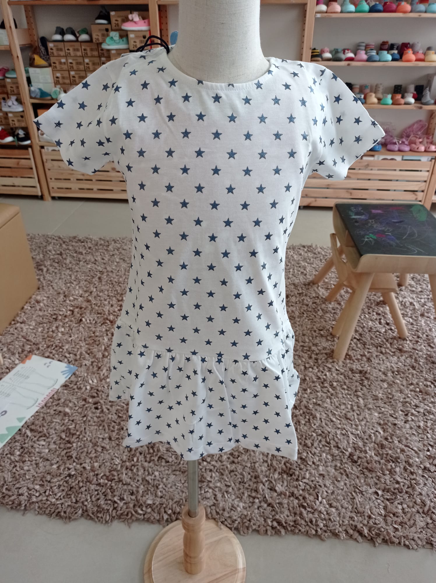 Vestido infantil Hublot branco com estrelas azuis, manga curta, em manequim, mostrando o corte com folho na bainha.