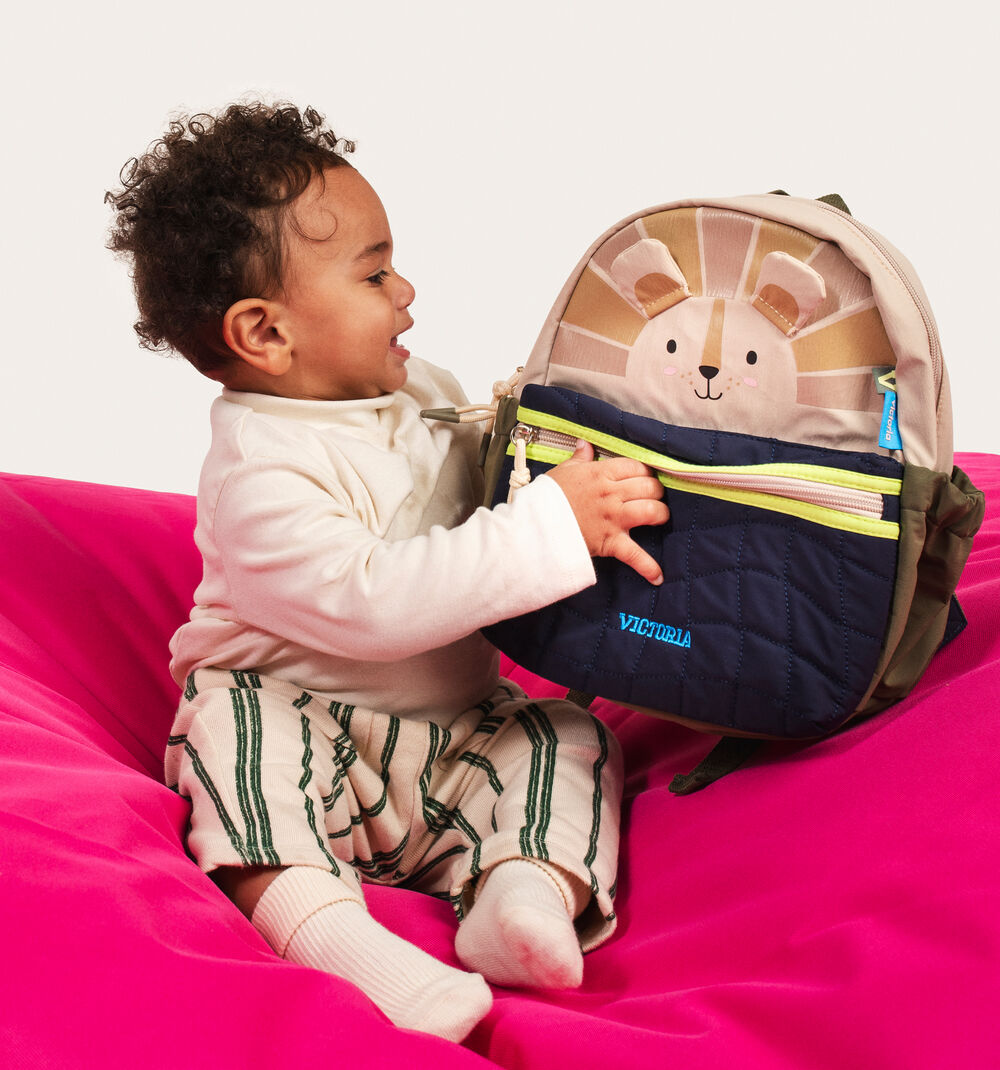 Bebé sorridente sentado num pufe rosa a interagir com a mochila infantil Victoria com tema de leão.