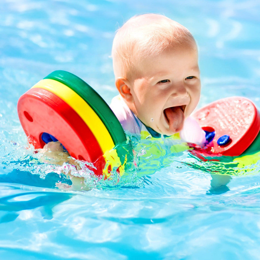 Um bebé feliz a sorrir e a salpicar água na piscina, a usar as braçadeiras flutuador Delphin Discs coloridas nos braços, desfrutando da água.