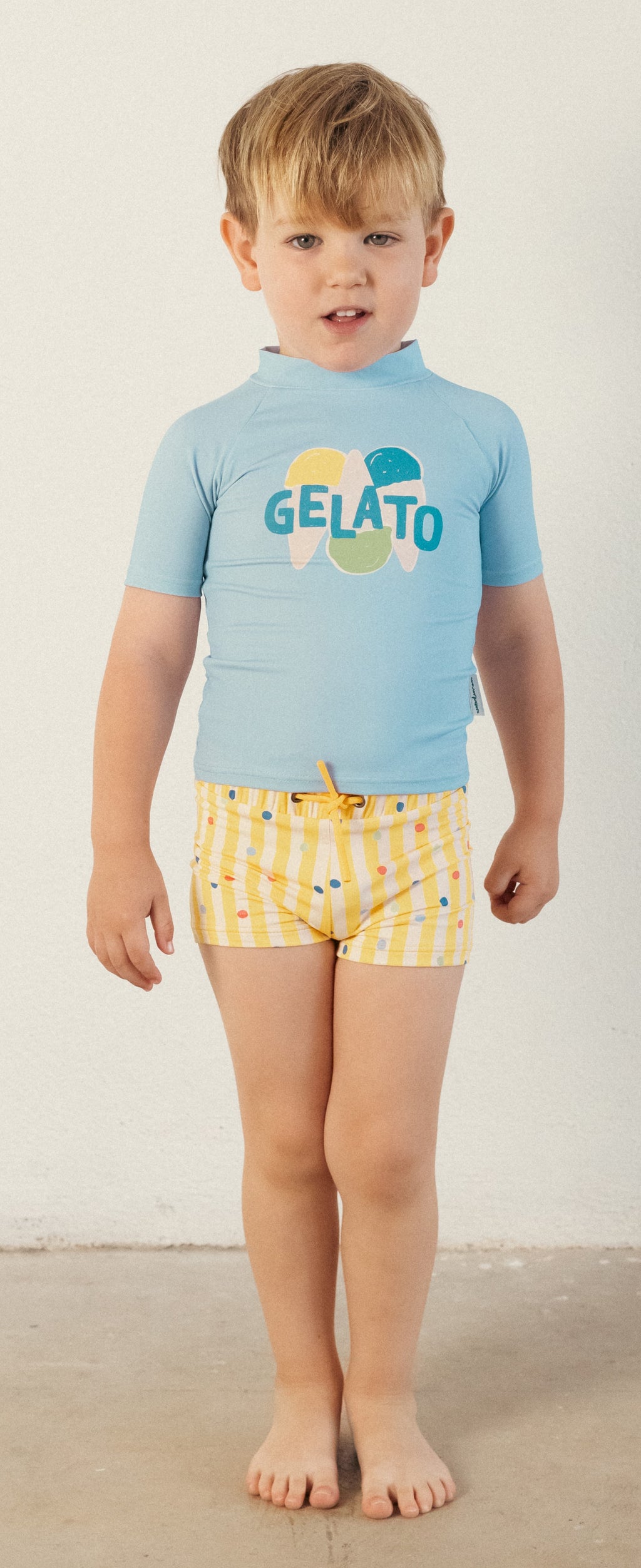 Criança a usar a boxer de praia amarela com riscas e bolinhas, combinada com uma camisola anti-UV azul clara com o desenho "GELATO".