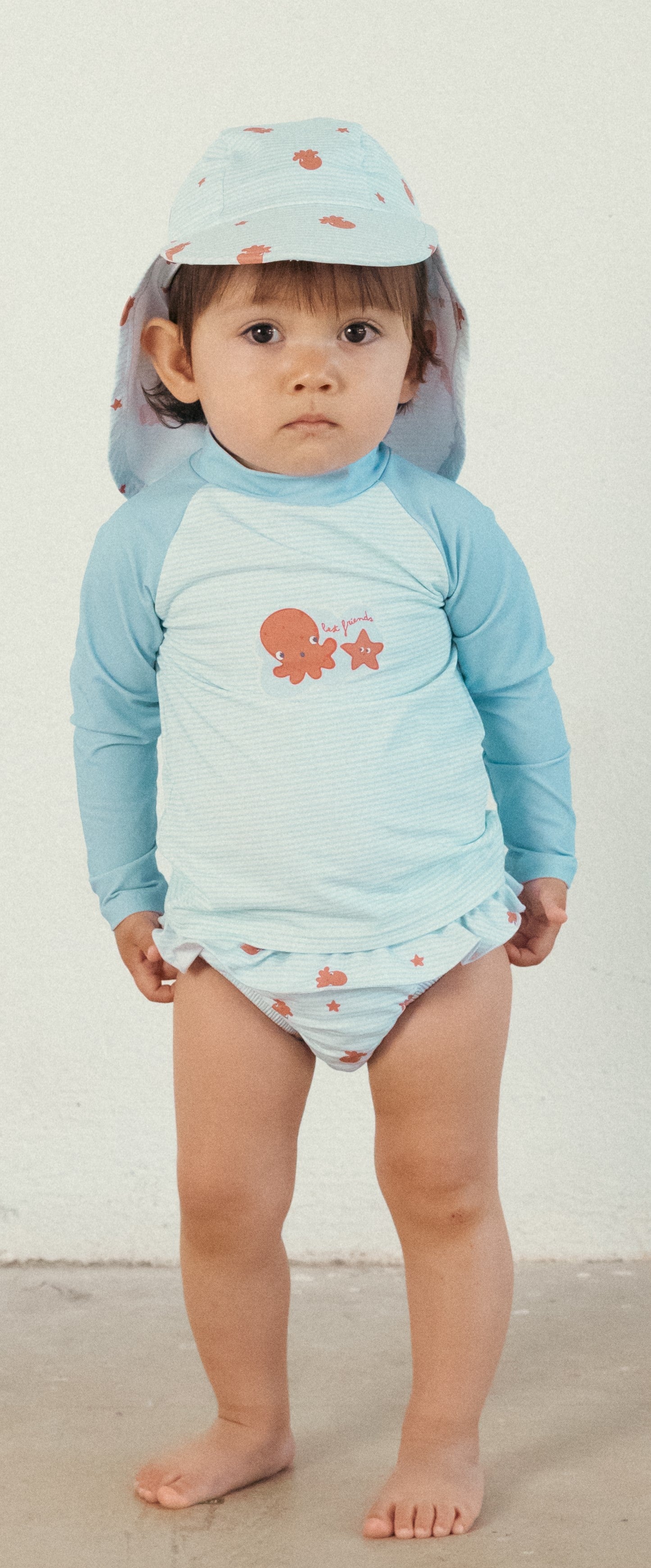 Bebé a usar a camisola de manga comprida UV Waterlemon com proteção solar, calções de banho e chapéu, em pose frontal.