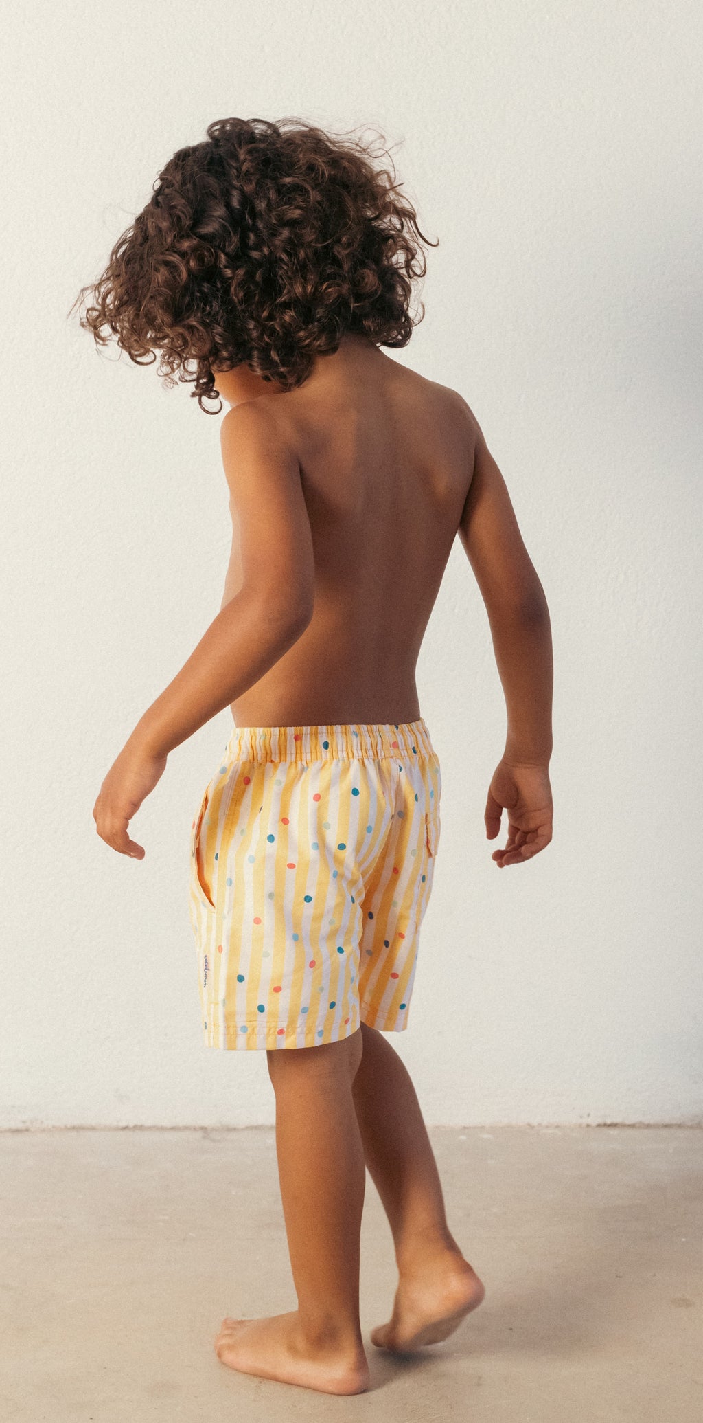 Criança vista de costas a usar o calção de praia Waterlemon amarelo, com cabelo encaracolado, em fundo claro.