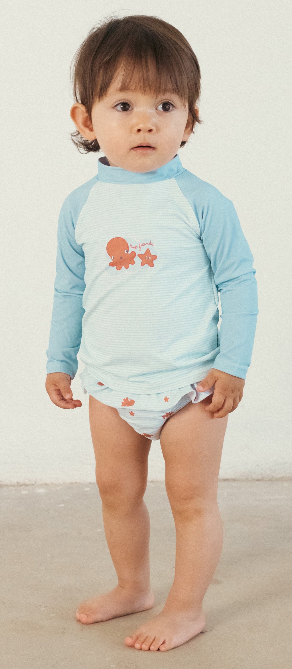 Bebé em pé a usar a cueca/fralda de praia Waterlemon azul com polvos e uma camisola de manga comprida clara.