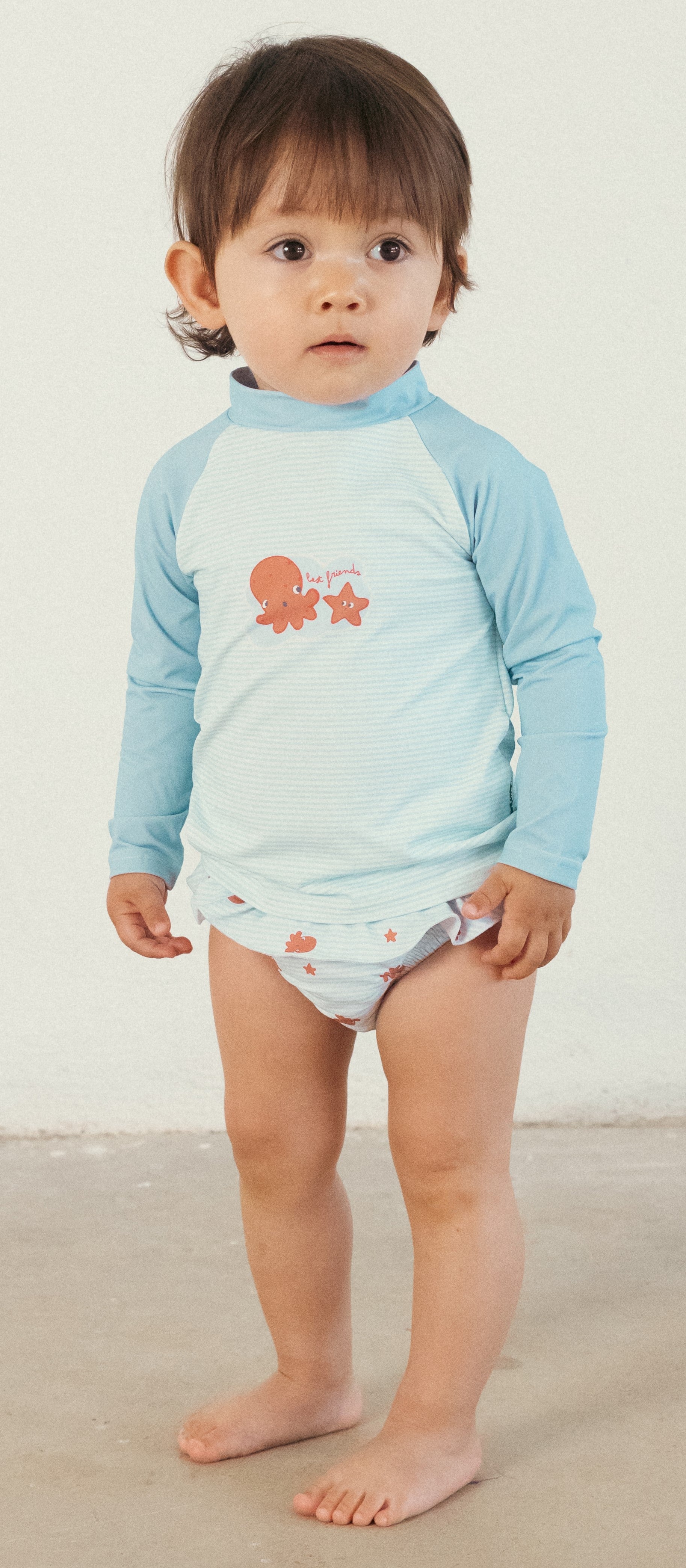 Bebé em pé a usar a cueca/fralda de praia Waterlemon azul com polvos e uma camisola de manga comprida clara.