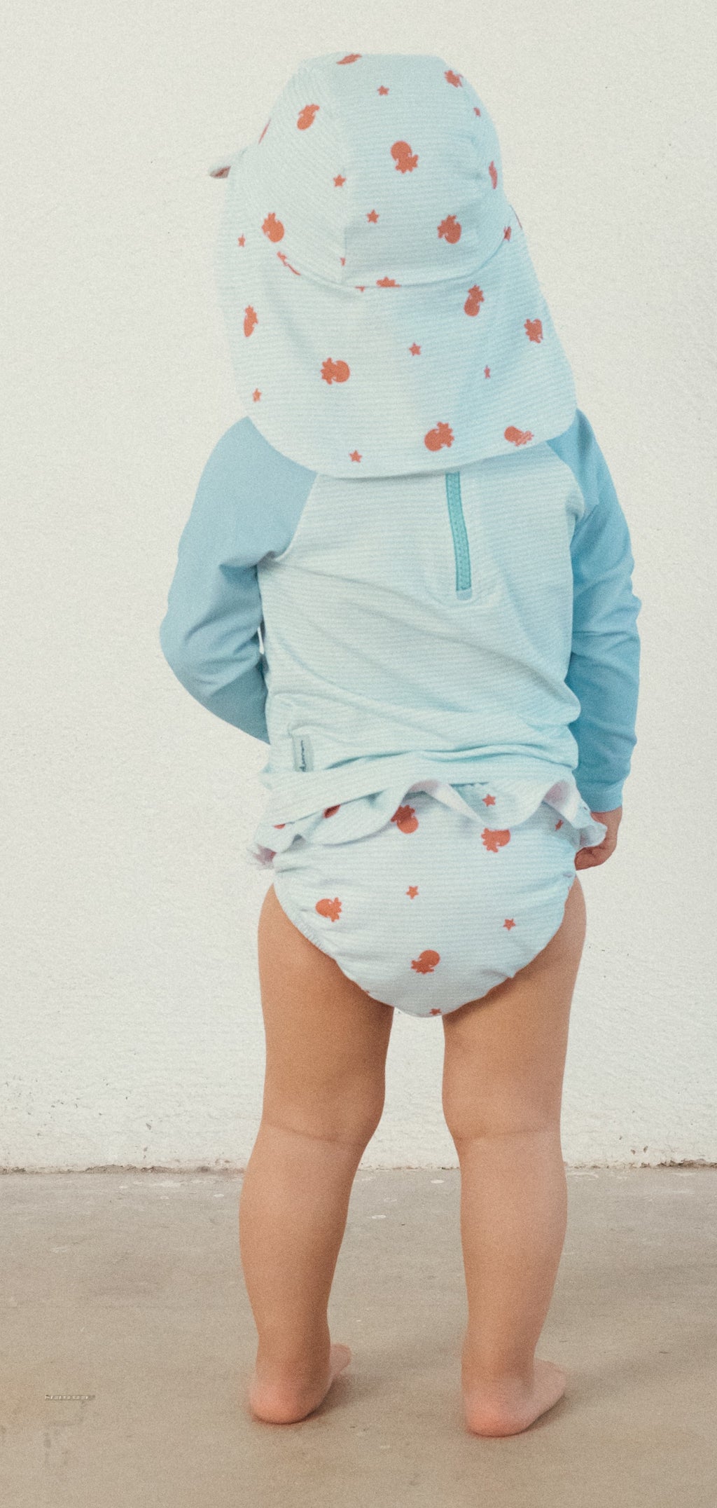 Vista traseira de um bebé de pé a usar a cueca/fralda de praia Waterlemon com folhos e um chapéu de sol a condizer.