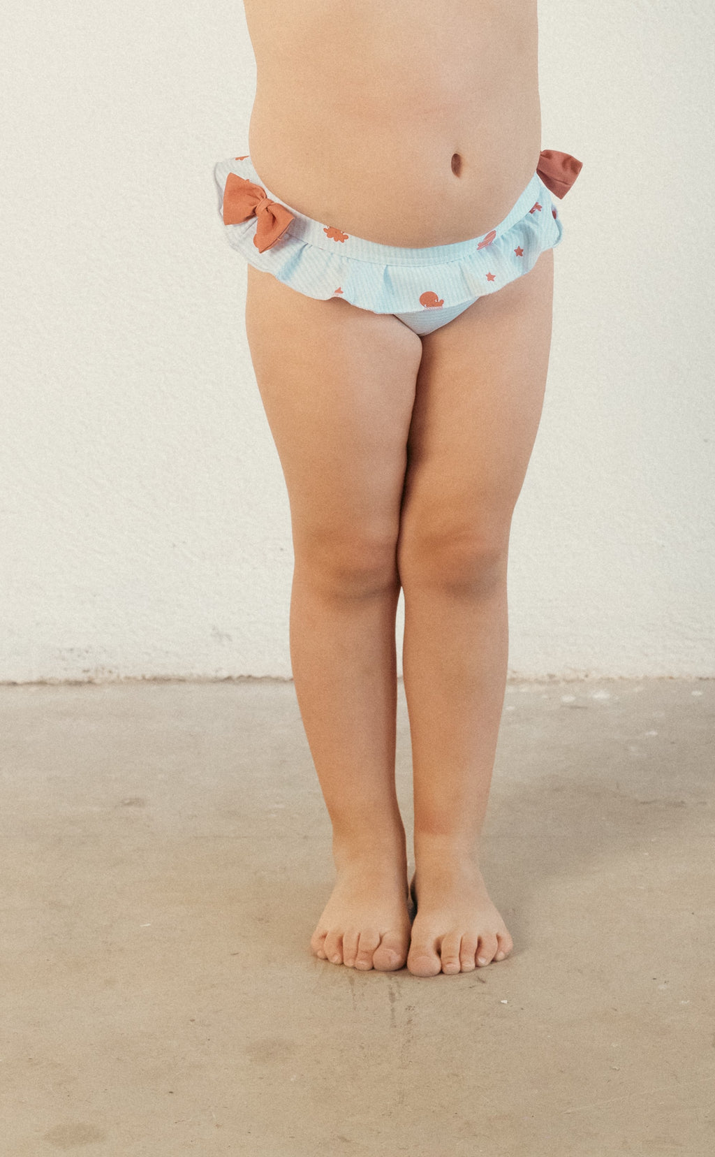 Criança em pé, vista frontal, a usar a cueca de banho com folhos Waterlemon, mostrando o ajuste confortável na barriga e pernas.