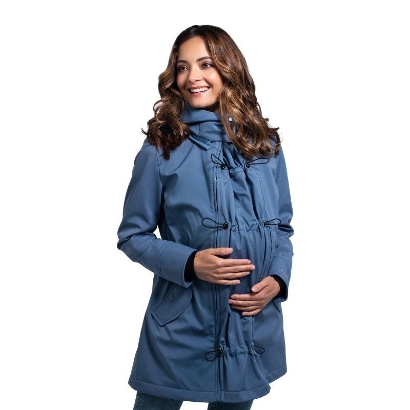 Mulher grávida sorridente com casaco Wombat Shell azul, mostrando o painel de expansão para a barriga.