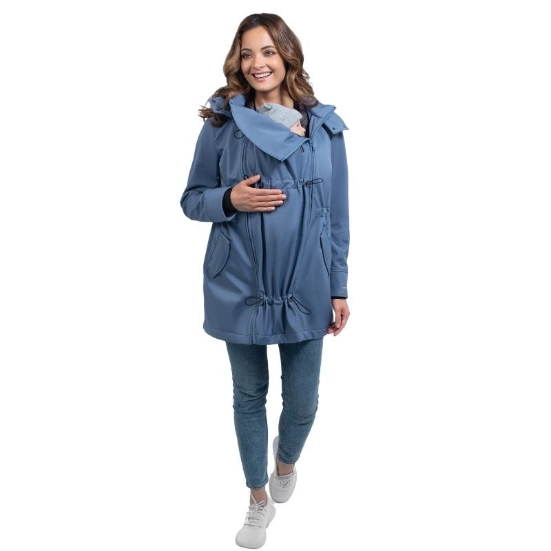 Mulher sorridente a caminhar com casaco Wombat Shell azul, usando o painel de transporte com o bebé oculto.