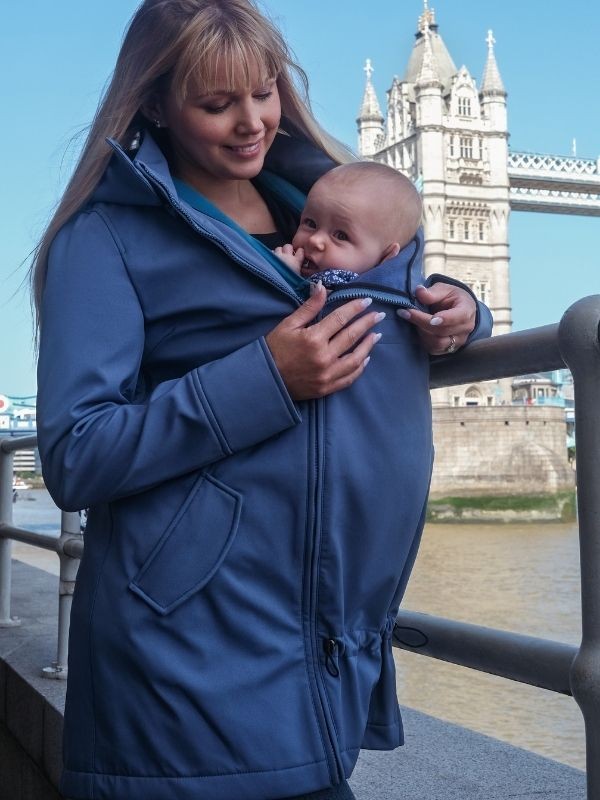 Mãe sorridente com casaco Wombat Shell azul a transportar um bebé à frente, com a Tower Bridge de Londres ao fundo.