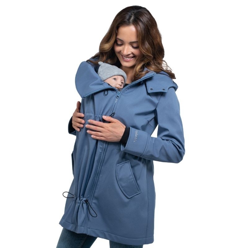 Mulher sorridente com casaco Wombat Shell azul a usar o painel de transporte com um bebé pequeno de gorro cinzento.