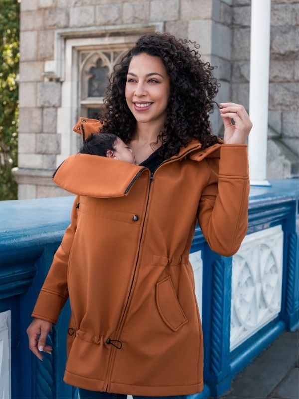 Mulher com cabelo encaracolado sorri ao usar o casaco Wombat Shell caramelo com um bebé a dormir no painel de transporte frontal.