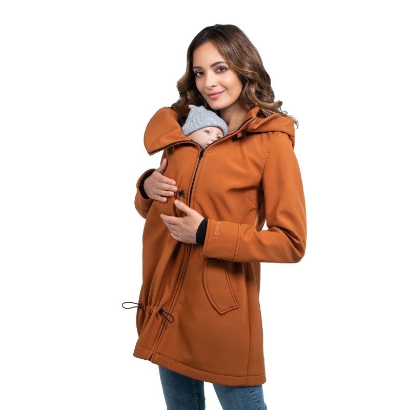 Mulher sorridente com o casaco Wombat Shell cor caramelo a usar o painel para transportar um bebé com gorro cinzento.
