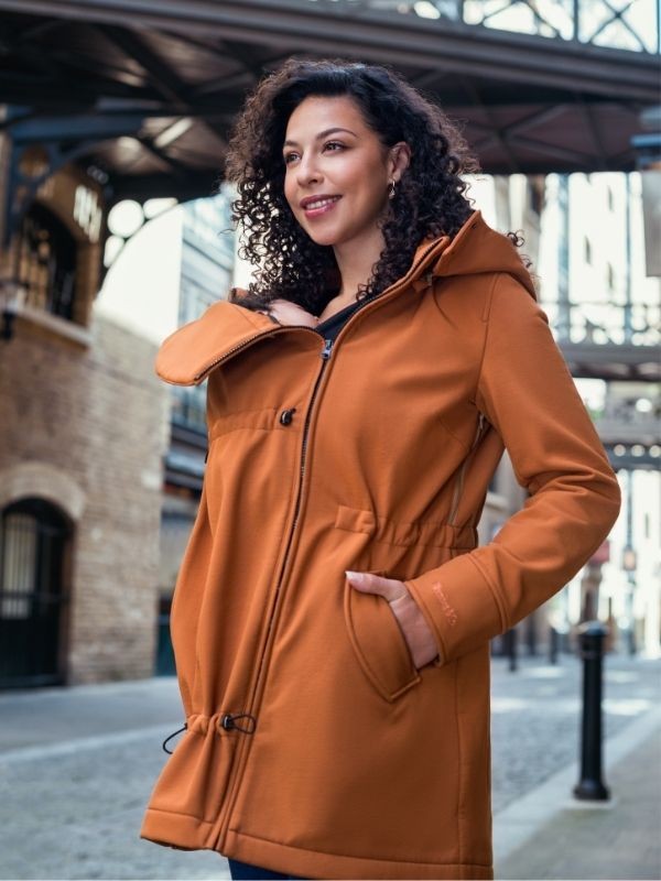 Mulher sorridente com casaco Wombat Shell caramelo, com um painel ajustado, num cenário urbano com arquitetura antiga.