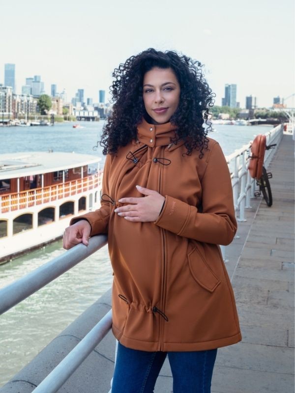 Mulher grávida sorridente com casaco Wombat Shell caramelo, usando o painel de expansão, junto a um cais com vista para a cidade.
