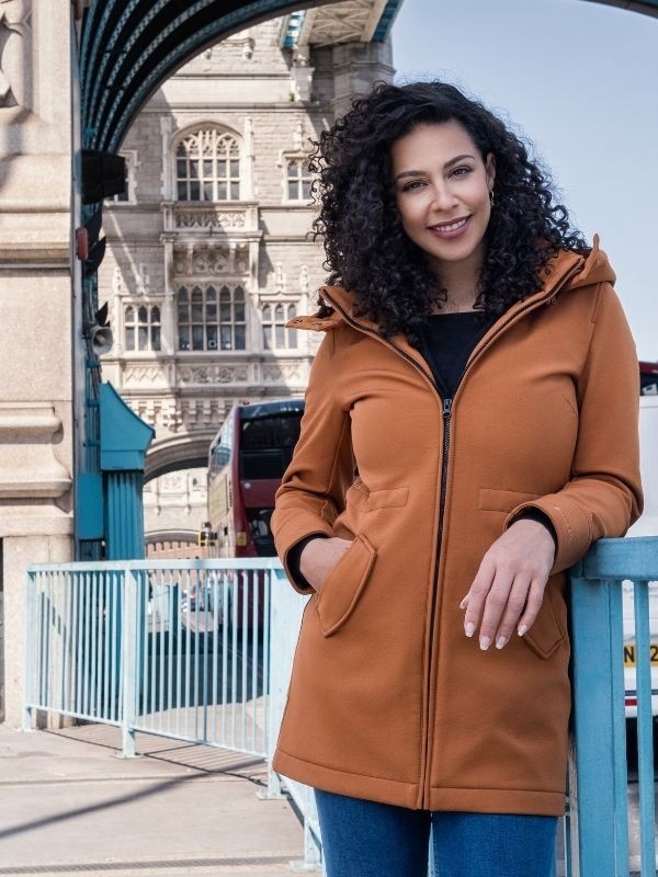 Mulher sorridente com casaco Wombat Shell caramelo, sem painel, em frente à Tower Bridge em Londres, estilo casual.
