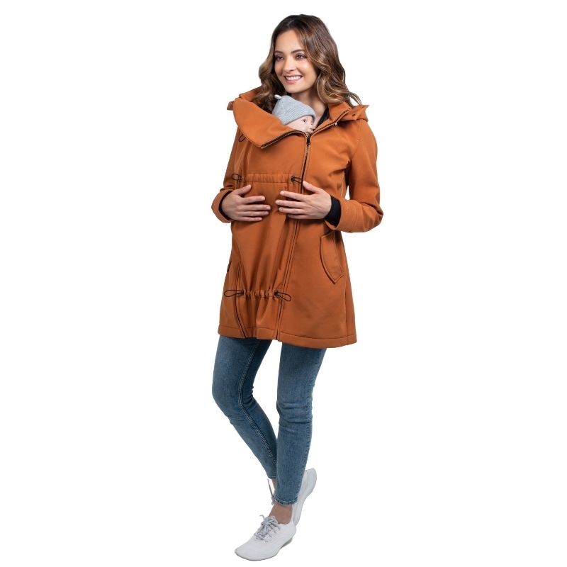 Mulher sorri usando o casaco Wombat Shell caramelo com um bebé pequeno transportado à frente, em fundo branco.