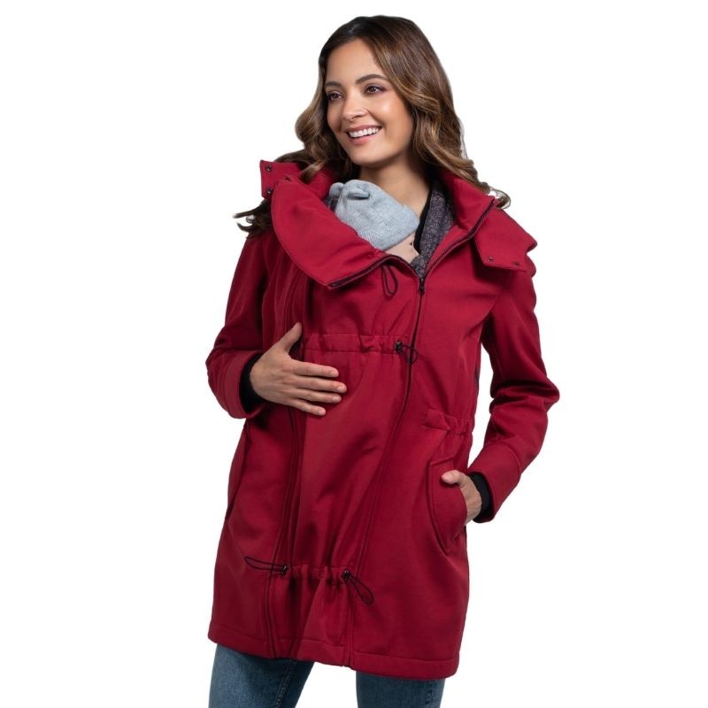 Mulher sorridente a usar o casaco Wombat Shell vermelho com painel de transporte frontal para bebé.