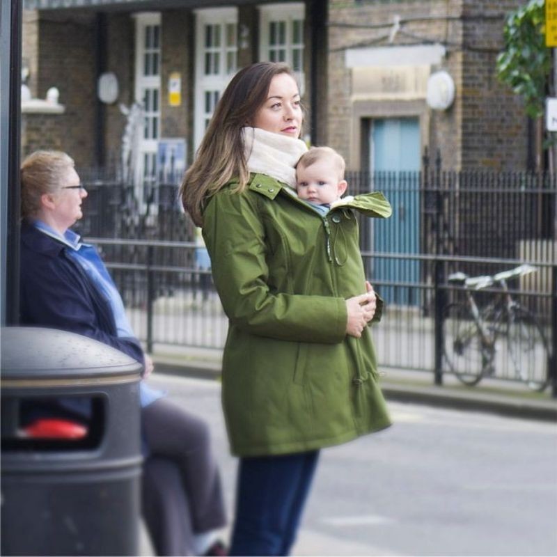 Mãe com casaco Wombat Wallaby verde a transportar um bebé à frente num exterior urbano, com inserção de transporte visível.