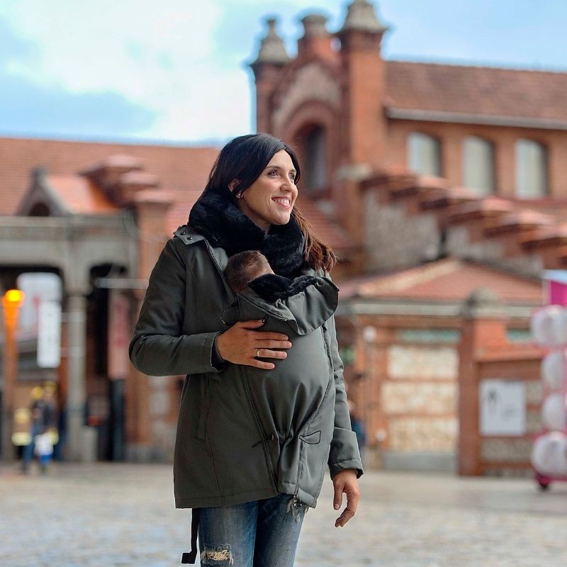 Mulher sorridente a usar o casaco Wombat Wallaby cinzento com o bebé no transporte frontal, ao ar livre numa rua urbana com edifícios de tijolo.
