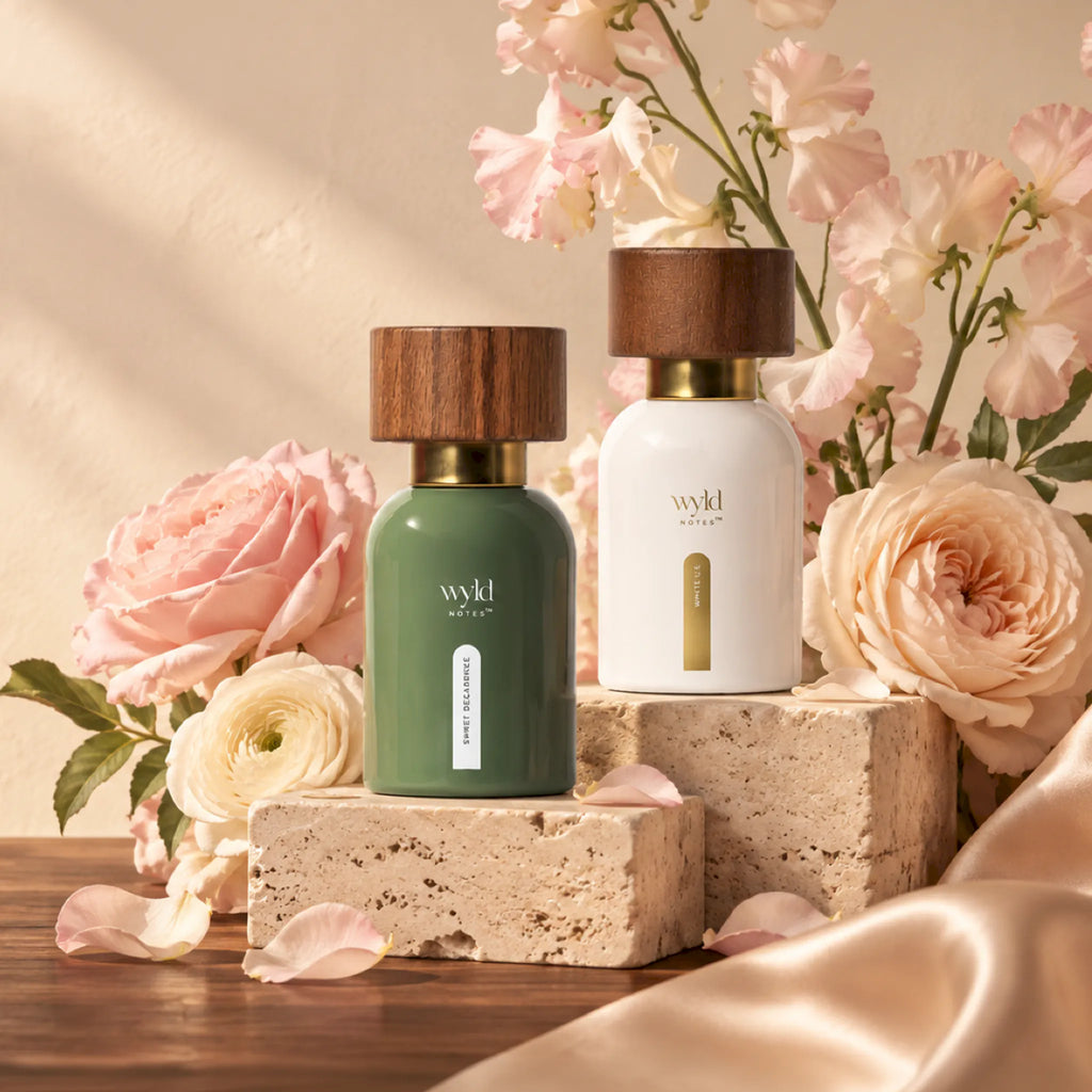 Os dois perfumes do Her Signature Duo pousados em pedra clara, rodeados de rosas e seda, em luz natural quente