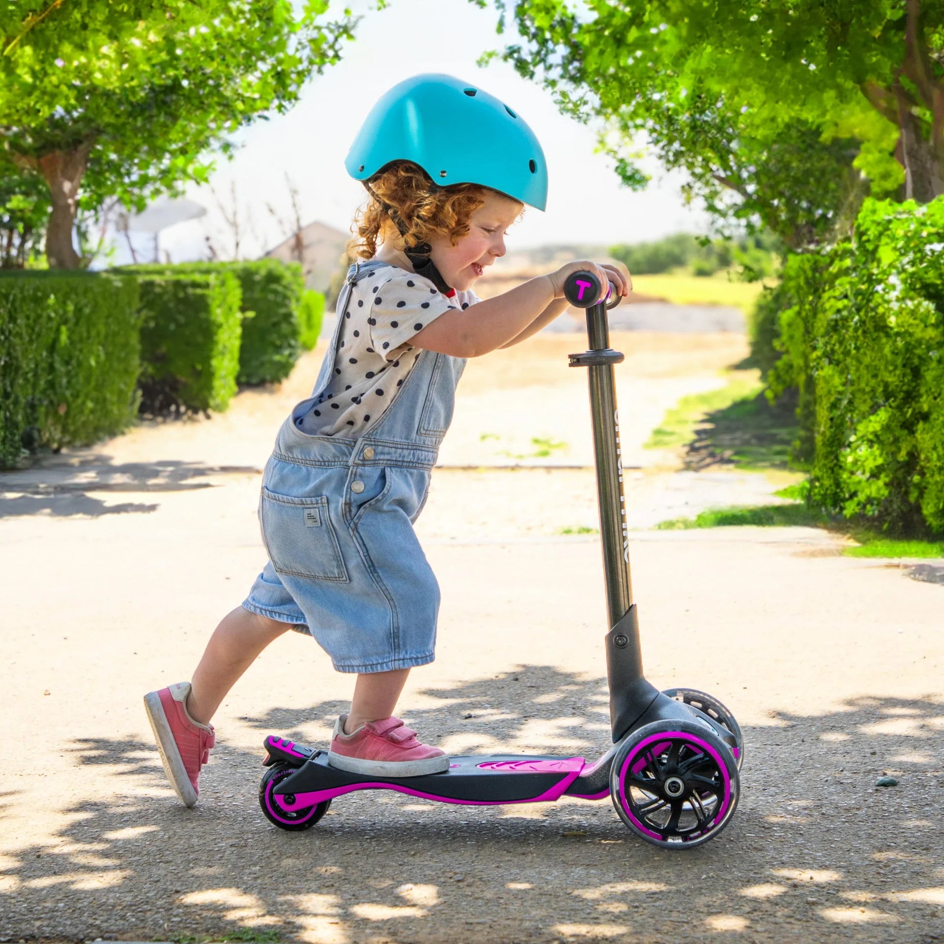 Criança a andar de Xtend Scooter Pink, um trotinete infantil ajustável em preto e rosa, ideal para desenvolver equilíbrio e coordenação. A criança usa um capacete azul para segurança e desliza num ambiente ao ar livre, rodeada de árvores e natureza.

