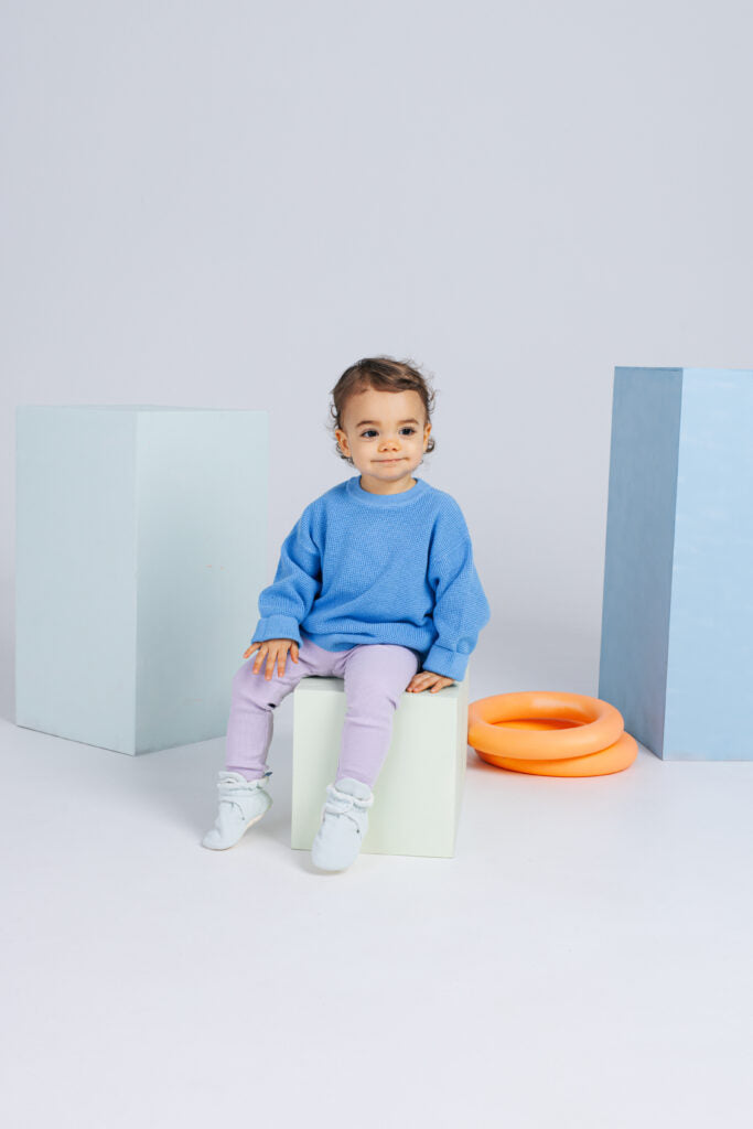 Bebé sorridente sentado num bloco, vestindo camisola azul e leggings lilases, com as pantufas botinhas Zás Trás Ice Shake nos pés.