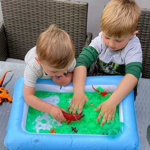 Crianças a explorar a textura sensorial do Gelli verde com as mãos, interagindo com as figuras de dinossauros na bandeja insuflável azul.