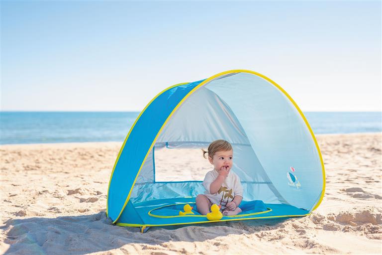 Bebé sentado dentro da Olmitos Tenda Piscina Pop Up azul e amarela na praia, com o mar ao fundo.