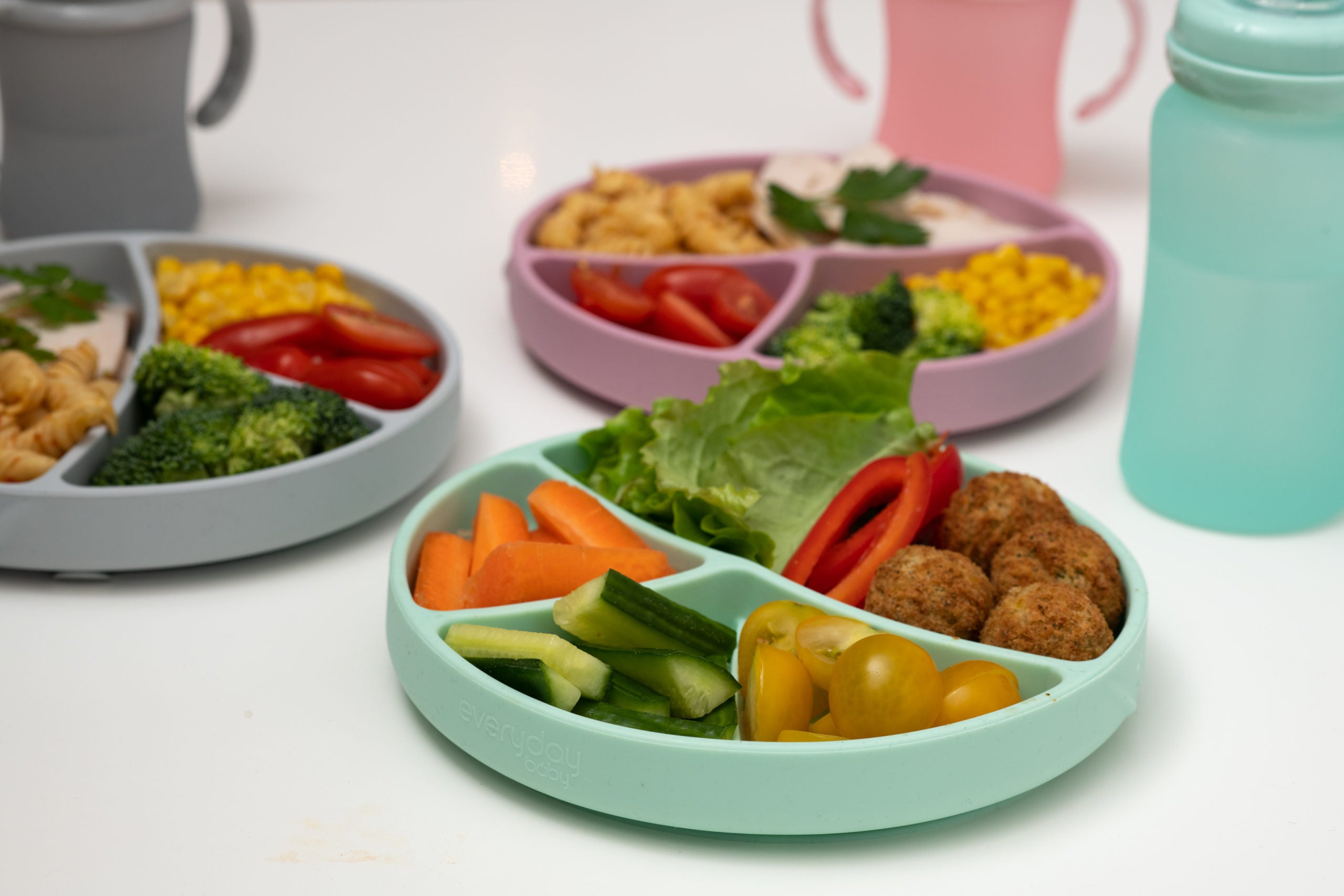 Três pratos de silicone da Everyday Baby, um cinzento, um rosa e um verde-água, cheios de comida variada como massa, vegetais e carne, sobre uma superfície branca.