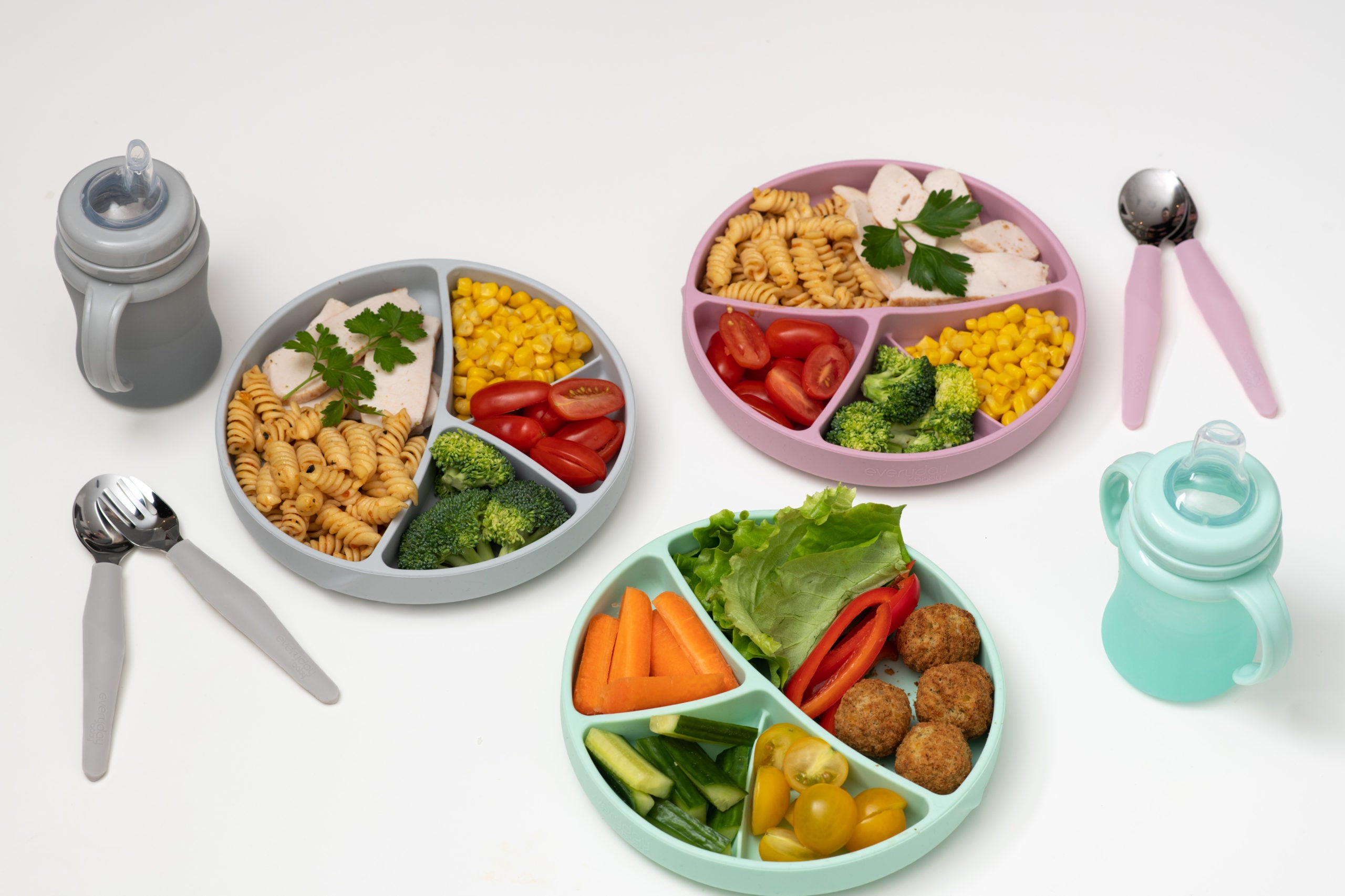 Três pratos de silicone compartimentados com refeições completas, rodeados por dois conjuntos de talheres e dois biberões, demonstrando o uso dos produtos na hora da refeição.