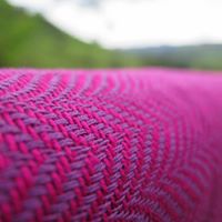 Imagem de um pano tecido Cazulim T6 Pafisae com uma trama chevron arredondada em tons de roxo, demonstrando a textura fina e a qualidade do tecido sob um fundo desfocado de paisagem verde.