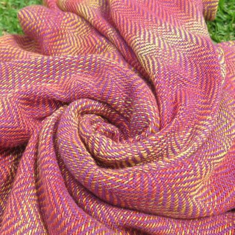 Imagem de um pano tecido Cazulim T6 Hera em close-up, mostrando uma trama chevron arredondada em tons de roxo e um degradê de laranja/amarelo, enrolado de forma a exibir a textura e cores vibrantes do tecido.
