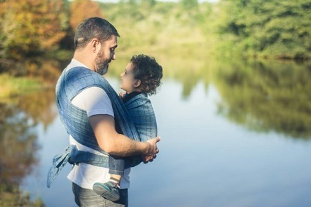 Homem segurando uma criança num pano tecido azul, em pé à beira de um lago, com árvores ao fundo. Ambos estão sorridentes e relaxados, aproveitando a tranquilidade do ambiente natural.