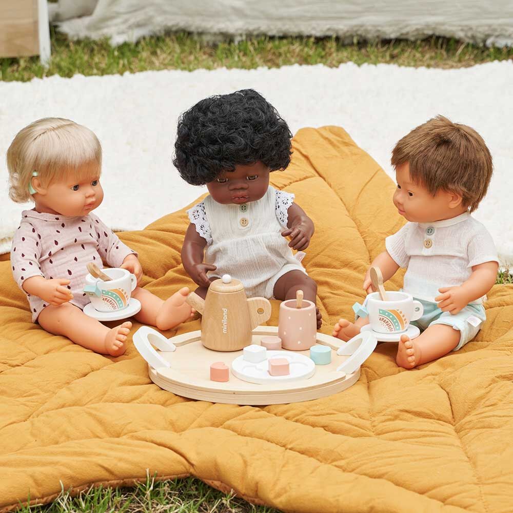 Três bonecos Miniland de diferentes etnias (caucasiano, africano, com Síndrome de Down) sentados num cobertor amarelo a brincar com um serviço de chá de madeira.