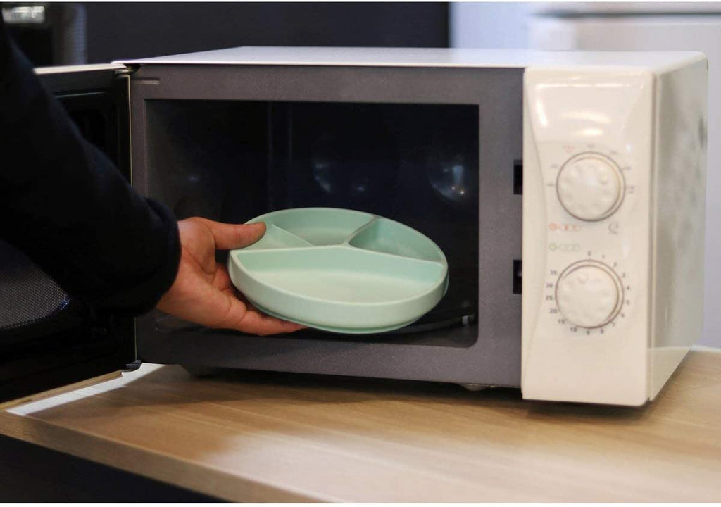Mão a colocar um prato de silicone verde-água com divisórias dentro de um micro-ondas branco.
