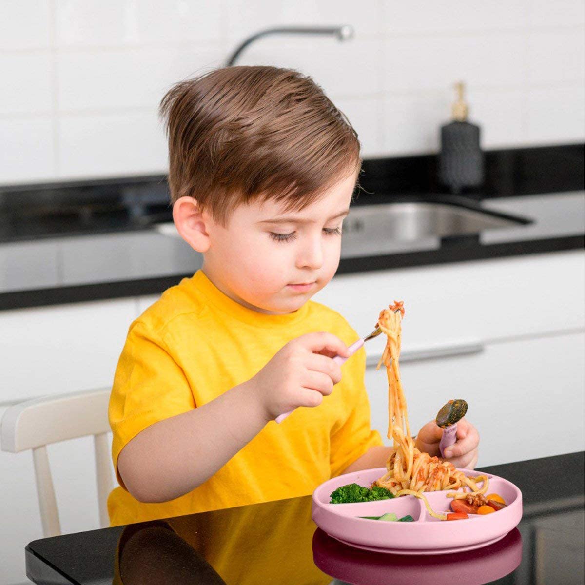 Rapaz a comer esparguete com um garfo a partir de um prato de silicone rosa com divisórias, sentado numa cozinha moderna.