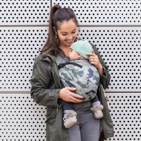 Mulher com cabelo escuro em um casaco, segurando um bebé num sling camuflado, demonstrando o uso do sling em ambientes urbanos.