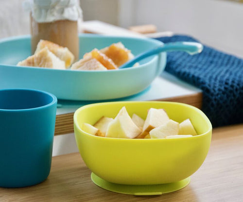 Conjunto de alimentação Beaba de 4 peças em silicone turquesa e verde lima, com prato, taça, chávena e colher, contendo fruta cortada e um frasco com comida para bebé, sobre uma mesa de madeira.