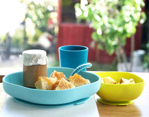 Vista aérea de um conjunto Beaba de alimentação em silicone turquesa e verde lima, com prato, taça, chávena e colher, com comida, junto a uma janela com plantas.