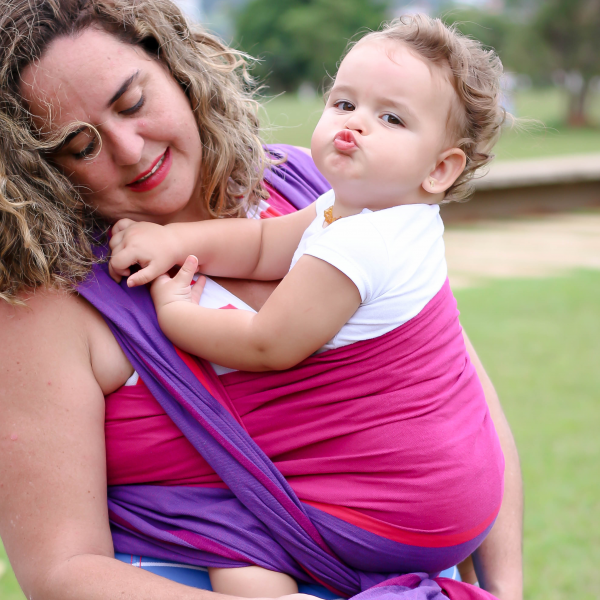 Uma mãe sorridente, com cabelo castanho e ondulado, usa um pano tecido cor de rosa e roxo para carregar o seu bebé. O bebé, de pele clara e cabelo castanho, está posicionado de frente para a mãe e faz uma expressão engraçada com os lábios. Ambos estão ao ar livre num espaço verde, transmitindo uma sensação de alegria e ligação.