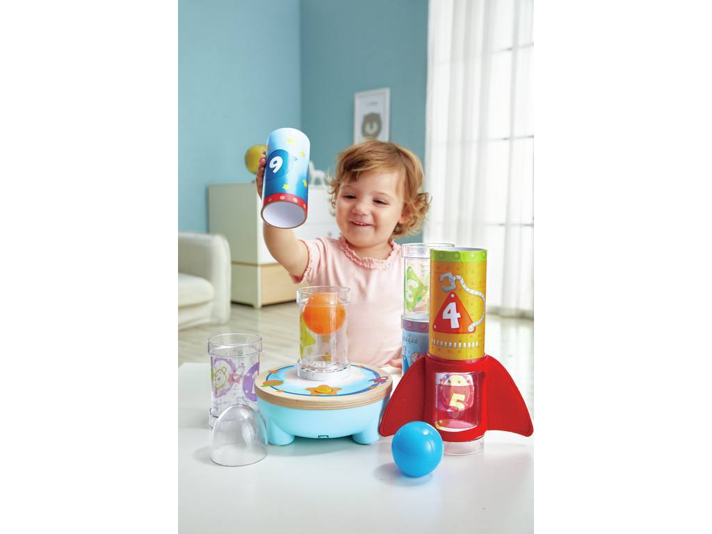 Uma menina sorridente a brincar com o Hape Foguetão com Bola, segurando um dos tubos numerados e preparando-se para o empilhar na base.
